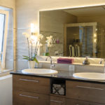 Badezimmer mit Natursteinwänden, einem großen Spiegel in der Mitte von 2 Waschbecken, eine Orchidee ist links neben dem linken Waschbecken